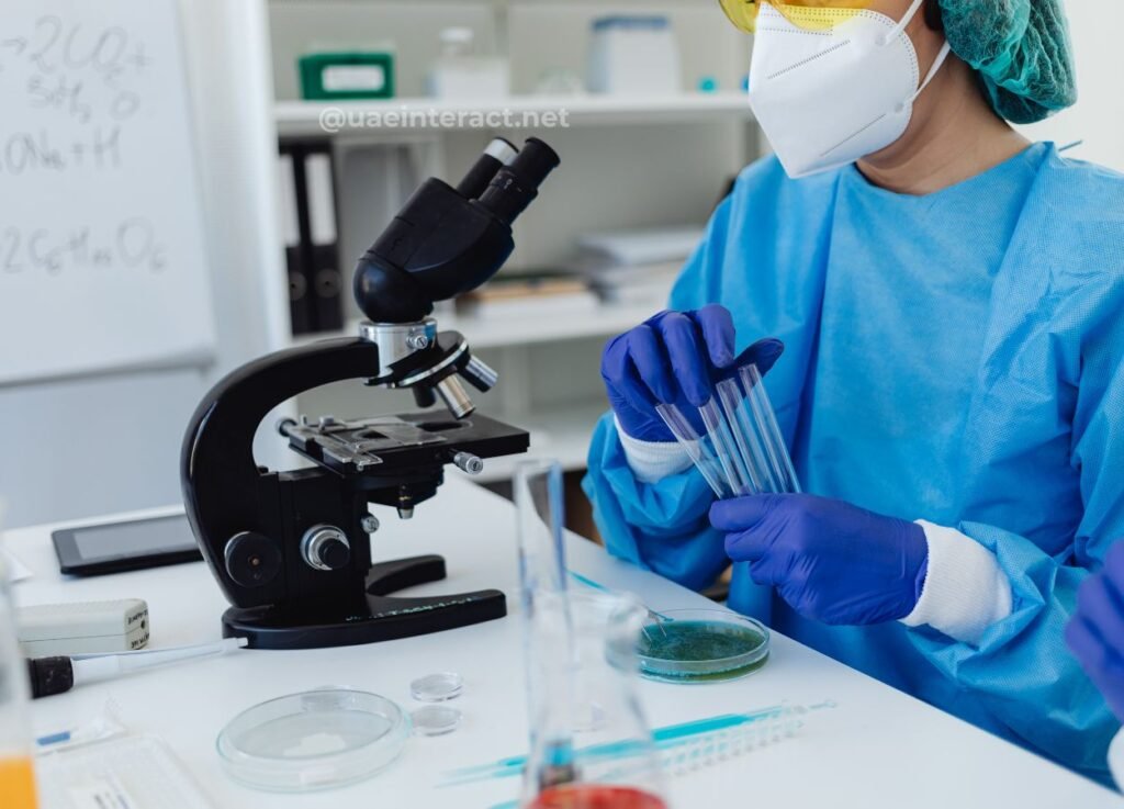 Scientist in PPE with mask and gloves holds clear test tubes beside a microscope in a lab environment.