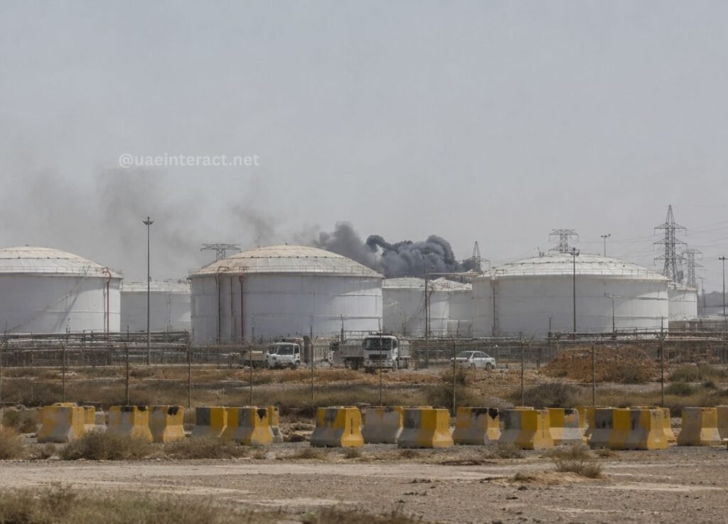 Large white cylindrical oil storage tanks at an industrial facility with dark smoke rising in the distance and vehicles near a fenced area in the foreground.