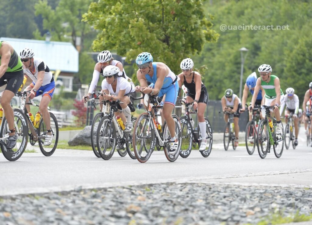 Group of cyclists in a road race, wearing colorful jerseys and helmets, riding closely on a paved street with green trees in the background.