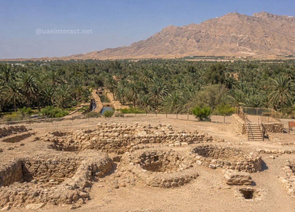Ancient stone ruins in a desert, with a palm oasis and rugged hills in the background.
