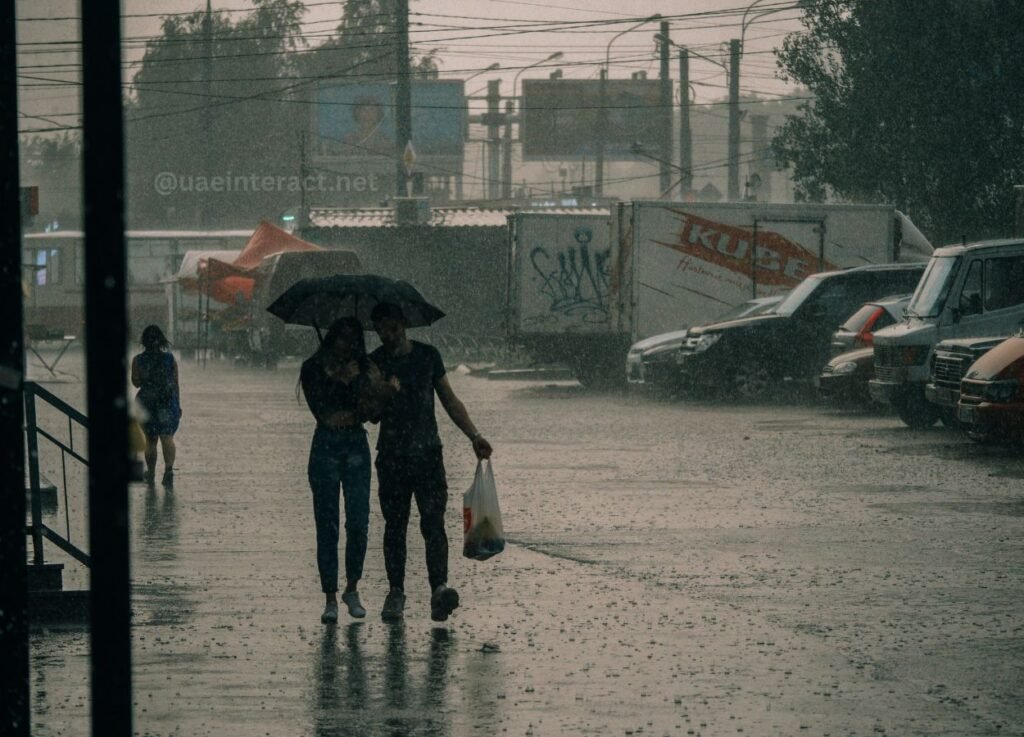 Couple walking under a dark umbrella in heavy rain on a wet urban street, with a pedestrian in blue in the background and graffiti-covered trailers nearby.
