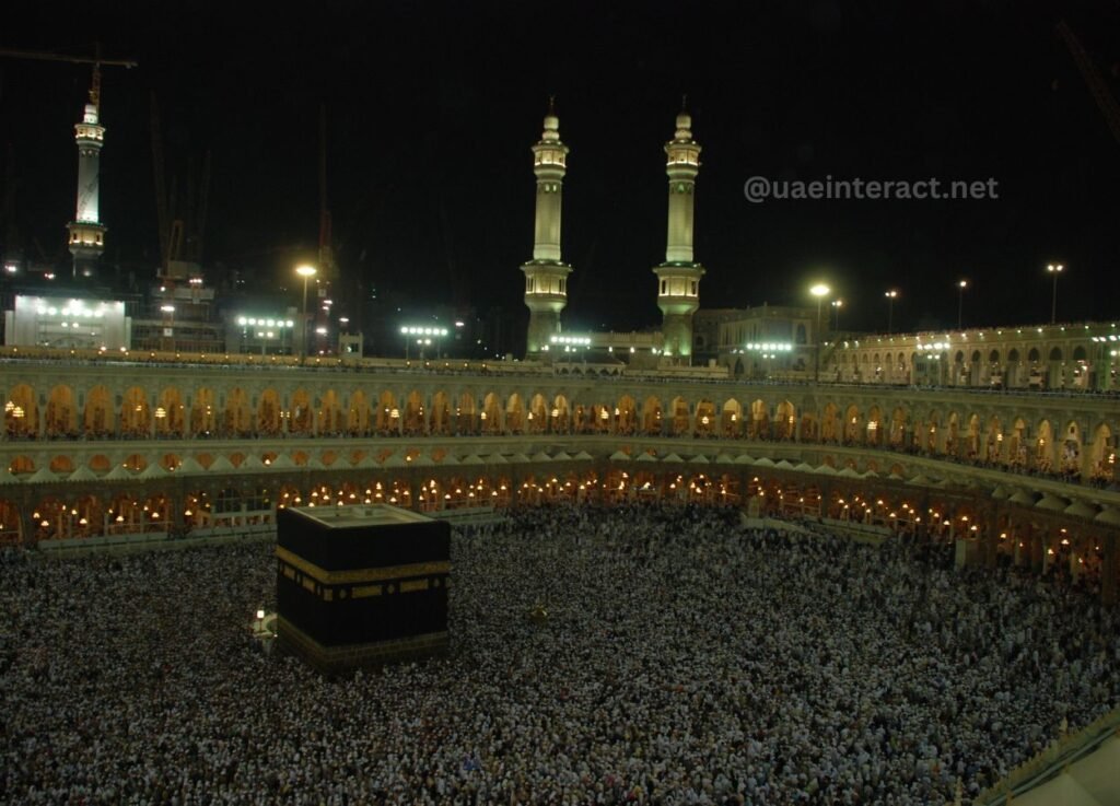 Night view of the Kaaba surrounded by a dense crowd at the Masjid al-Haram in Mecca, illuminated by numerous lights and minarets in the background