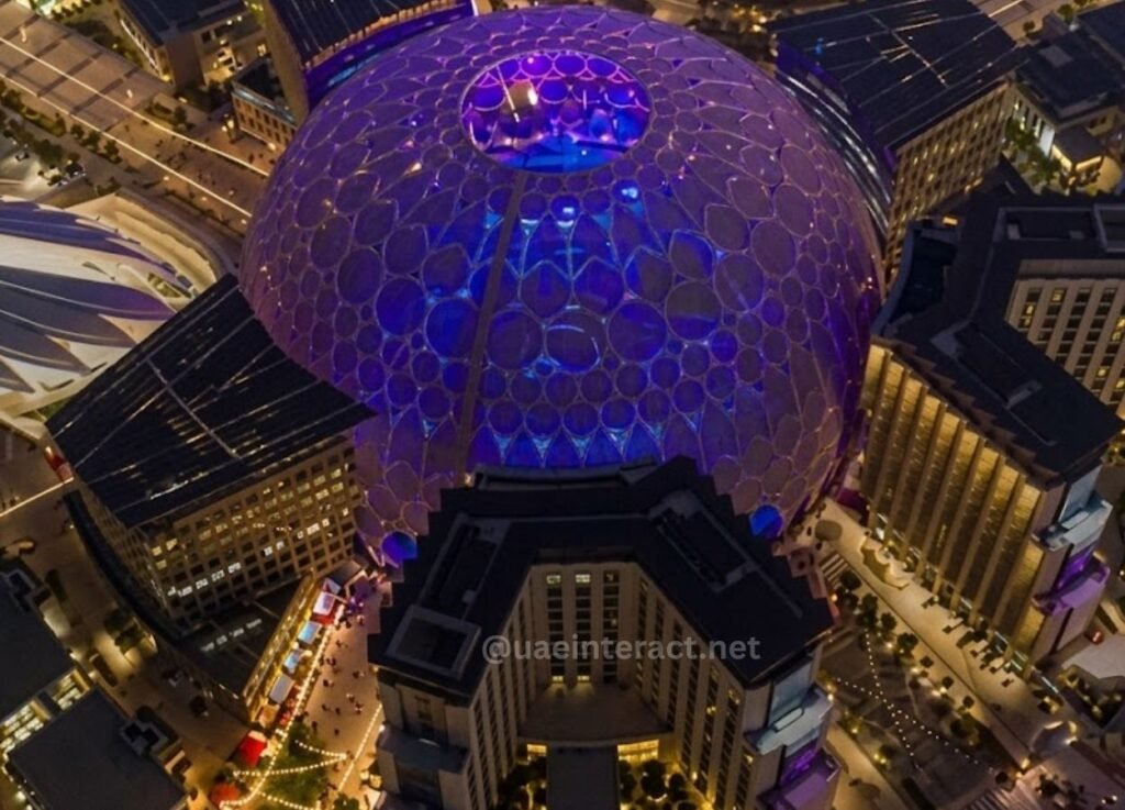 - UAE Interact Aerial night view of a large circular dome illuminated in purple, surrounded by tall city buildings and lit streets.