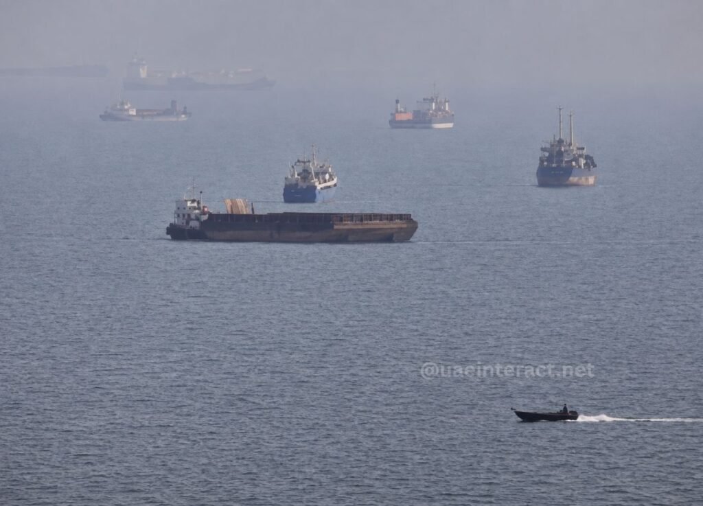 - UAE Interact A large cargo barge in the foreground on a calm sea, with several distant ships and a small speedboat speeding by on the right.
