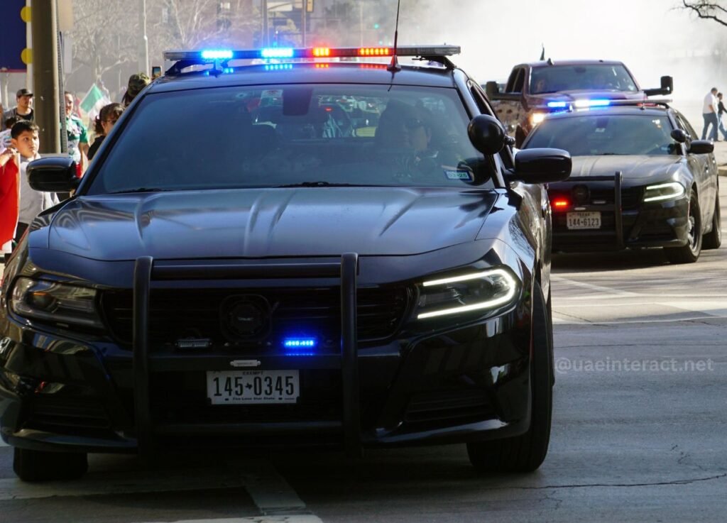 12 - UAE Interact Police SUVs with flashing blue lights leading a street procession as bystanders watch from the sidewalk.
