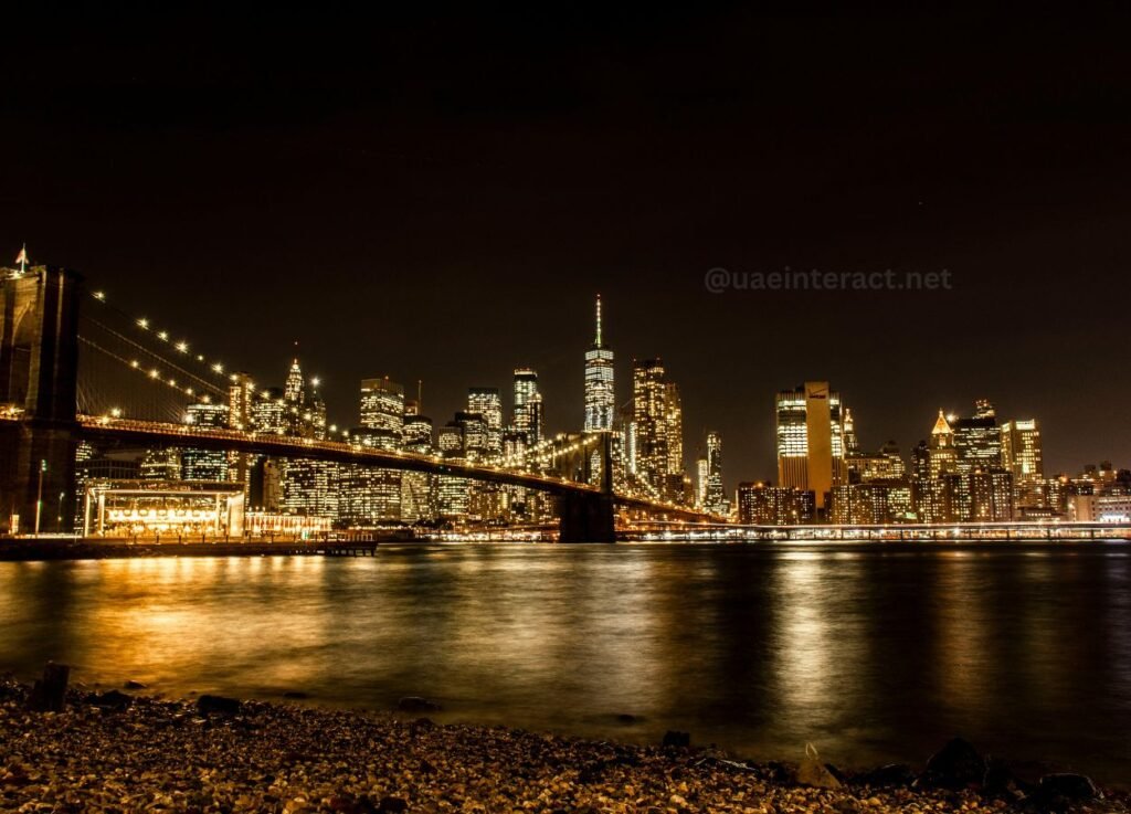 Nighttime city skyline with a lit suspension bridge spanning a river and reflecting lights on the water, with a rocky foreground shore.