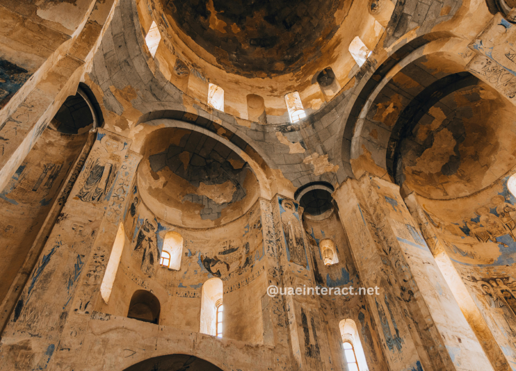 3 - UAE Interact Interior of an ancient domed church with tall arches and faded blue frescoes along the walls, lit by narrow arched windows