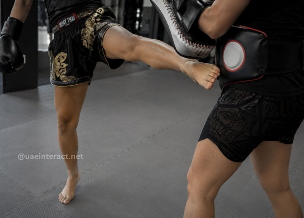 52 - UAE Interact Muay Thai practitioner delivering a kick to a padded target held by a partner in a gym, wearing ornate black shorts and gloves.