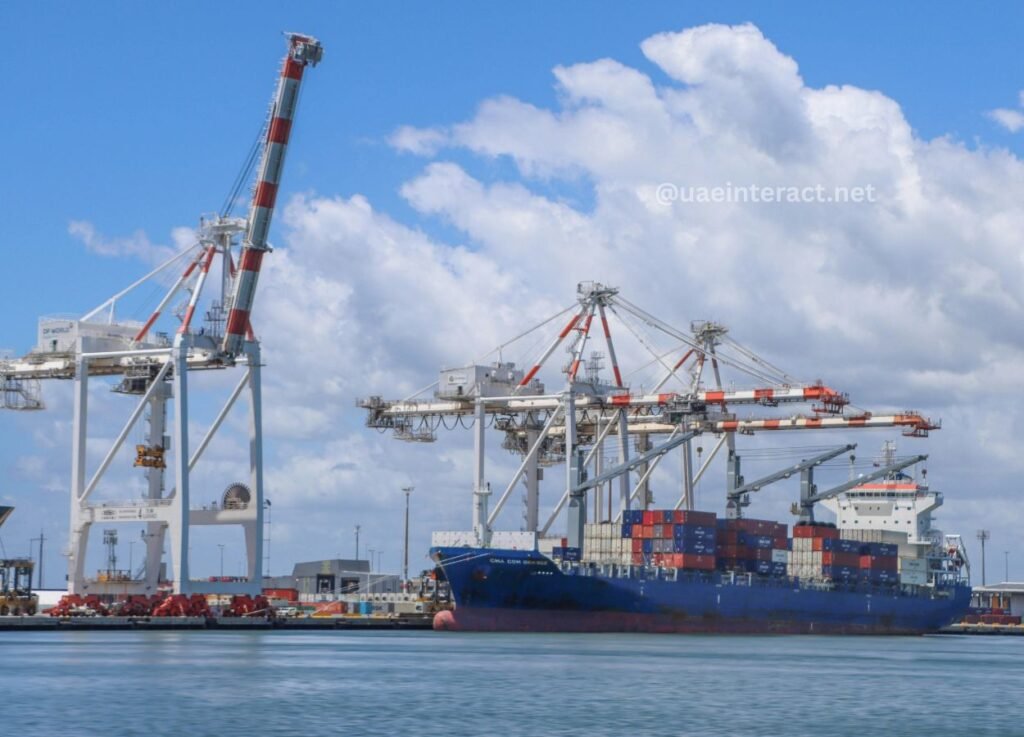 Blue container ship docked at a port with red-and-white gantry cranes unloading cargo containers under a bright sky.