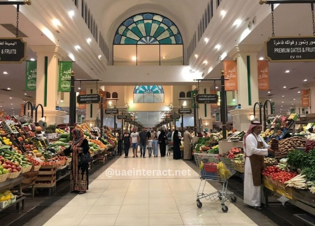 Indoor market aisle with fruit and vegetable stalls on both sides and shoppers browsing; man in traditional attire by a cart on the right.
