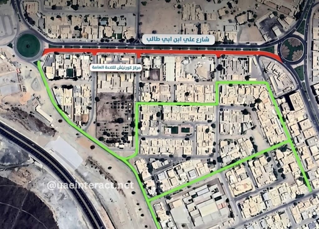 Aerial map of a desert city with a red main road and green detour routes, labeled in Arabic, showing roundabouts and blocks of buildings.